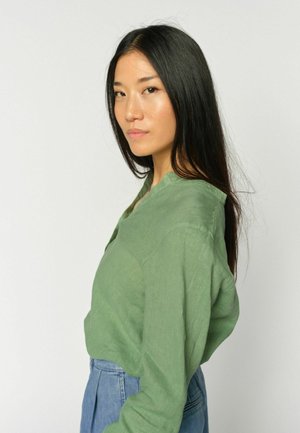 Junge Frau mit langen schwarzen Haaren, die eine grüne Bluse und blaue Jeans trägt und seitlich vor einem schlichten hellen Hintergrund blickt.