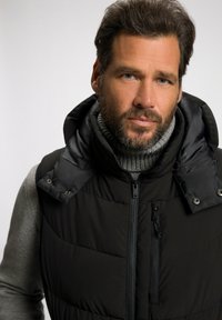 Schwarze Steppweste mit abnehmbarer Kapuze, mit durchgehendem Reißverschluss, seitlichen Taschen und einem grauen gerippten Rollkragenpullover darunter. Strukturierter Stoff.