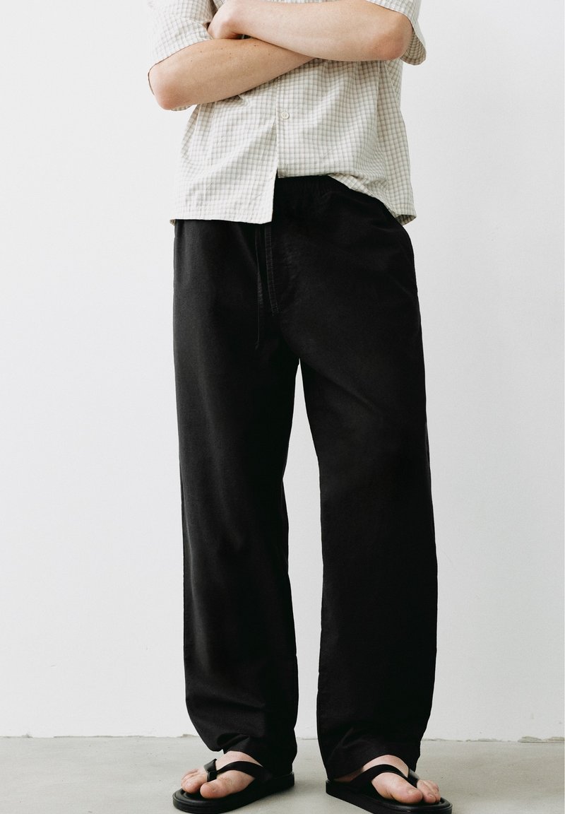 Personne portant un pantalon noir ample, une chemise à manches courtes beige à carreaux, les bras croisés, et des sandales noires, debout contre un mur blanc uni.