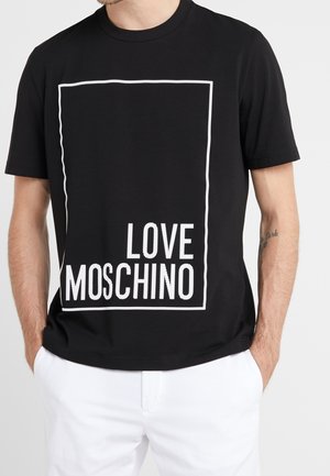 Mies, joka pukeutunut mustaan lyhythihaiseen T-paitaan, jossa valkoinen suorakaiteenmuotoinen reunus ja teksti "LOVE MOSCHINO", yhdistettynä valkoisiin housuihin, kädet taskuissa.