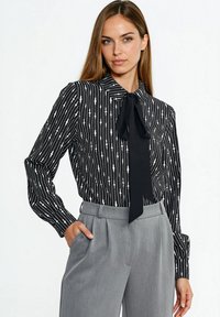 Bluzka w czarno-białe paski z kołnierzykiem i centralną kokardą; długie rękawy; geometryczne wzory; zestawiona z szarymi spodniami z wysokim stanem.