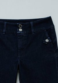 Jeans de mezclilla oscura con una textura suave, que presentan dos cierres de botón plateado y un acento decorativo en forma de diamante en el bolsillo.