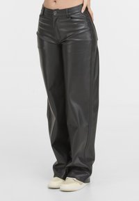 Pantalon en cuir noir taille haute à jambes droites porté avec des baskets décontractées beiges par une personne debout sur un fond uni.