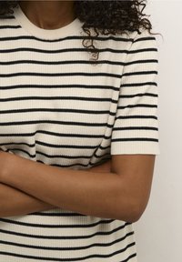 Haut à manches courtes crème avec rayures horizontales noires, texture côtelée et col rond. Bras croisés devant.