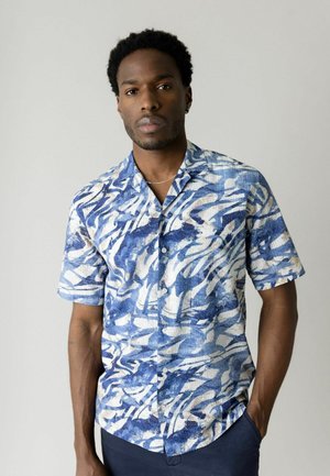 Mężczyzna o krótkich, kręconych włosach, ubrany w niebiesko-białą koszulę na guziki z abstrakcyjnym wzorem i krótkimi rękawami, stojący z jedną ręką w kieszeni.