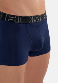 Námořnické modré trenýrky vyrobené z hladkého materiálu s černým elastickým pásem, na kterém je šedé logo. Obsahují přední kapsu a mají přiléhavý střih.