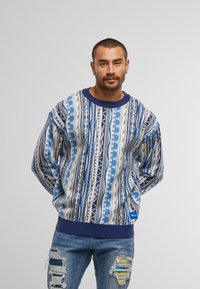 Patroon oversized trui in blauwtinten met geometrische ontwerpen, geribbelde manchetten en een blauwe label aan de onderkant. Gecombineerd met versleten spijkerbroek.