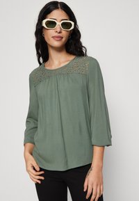 Grønn bluse med blonder ved bypassen, 3/4-ermer, flytende stoff og avslappet passform. Kombinert med hvite oversized solbriller.