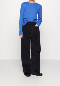 Modrá košile s dlouhým rukávem kombinovaná s volnými černými manšestrovými kalhotami. Košile má kulatý výstřih a kalhoty mají přední kapsy.