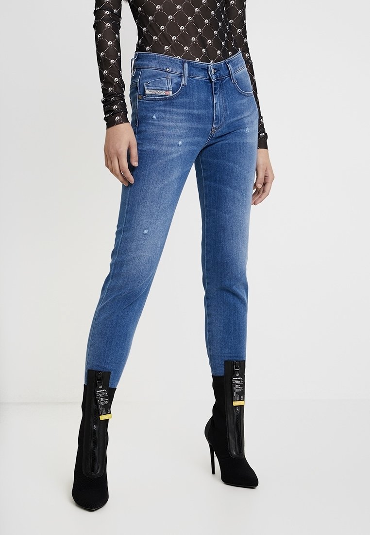 Jean en denim bleu à coupe slim, avec une taille mi-hauteur et des effets d'usure subtils. Associé à des bottines noires texturées à talons.