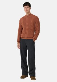 Pull en maille torsadée de couleur orange rouille, avec un col montant et des poignets côtelés. Associé à un pantalon sombre ajusté et des chaussures marron.