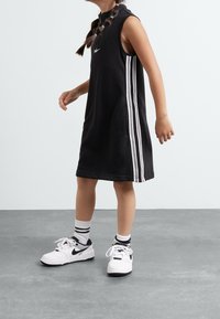 Vestido negro sin mangas con un cuello alto y un diseño de tres rayas blancas; combinado con zapatillas blancas y negras y calcetines con rayas negras.