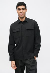 Mustast nuppudega särk kahe rinnataskuga, struktureeritud disainiga, pikkade varrukatega ja siledapinnaline. Kanditud mustade pükstega.