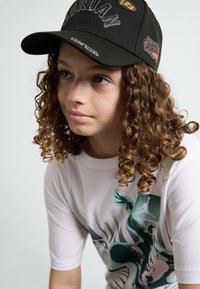 Svart caps med "Jordan"-logo og lapper. Hvit skjorte med grafisk design i grønt. Krøllete hår delvis synlig.