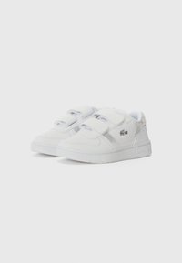 Baskets blanches avec des sangles Velcro, dessus en cuir, panneau latéral texturé, semelle en caoutchouc et un petit logo sur le côté.