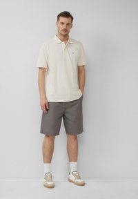 Polohemd aus leichtem beigem Baumwollstoff, kombiniert mit grauen Shorts. Das Modell trägt weiße Socken und Sneaker mit beigen Akzenten. Schlichtes Design, lässige Passform.
