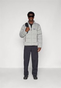 Mężczyzna w szarej kurtce puchowej, czarnych spodniach typu cargo, czarnych butach, dużych okularach przeciwsłonecznych, trzymający pasek czarnego plecaka na białym tle.