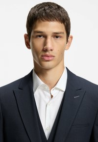 Veste de costume bleu marine foncé portée sur une chemise blanche à texture subtile ; fermeture par un bouton unique et petit détail rouge sur le revers.