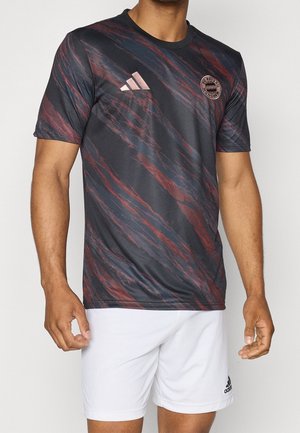 Mężczyzna ubrany w czarno-czerwoną, w paski, koszulkę FC Bayern München Adidas z krótkim rękawem oraz białe spodenki Adidas stojący na jednolitym tle.