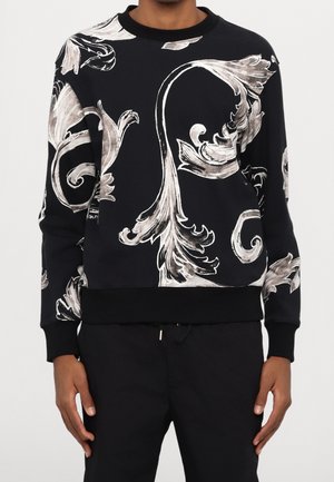 Zwarte longsleeve sweater met grote witte en grijze sierlijke bloemmotieven, gedragen met een zwarte broek.