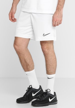 Mężczyzna w białych szortach Nike, białych skarpetkach Nike oraz czarnych butach sportowych Nike stojący na jasnoszarej podłodze.