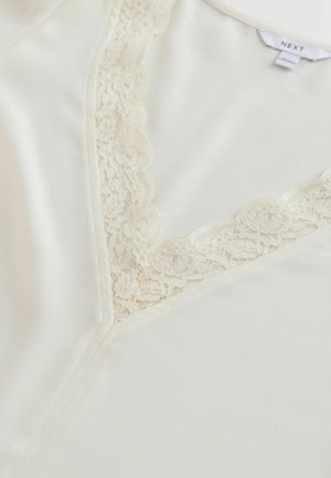 Cremefarvet bluse med V-hals, der har delikat blomsterlāc trim langs kanterne. Glat satinlignende tekstur, ingen synlige mønstre eller detaljer.