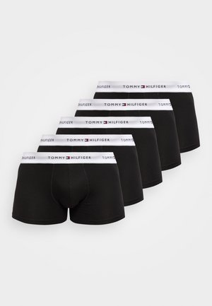 Homme athlétique debout pieds nus sur un tapis, portant un boxer noir avec une ceinture blanche Tommy Hilfiger, dans une pièce minimaliste avec un fauteuil blanc.