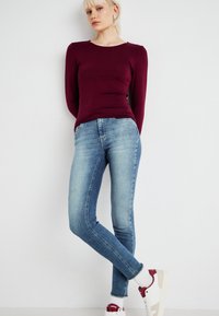 Burgunderfarget langermet tettsittende topp kombinert med lyseblå smale jeans. Modellen har på seg hvite joggesko med røde detaljer. Enkel design, uformell stil.