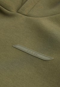 Felpa verde oliva realizzata in tessuto morbido, con un'etichetta cucita etichettata "CORE" in pelle verde chiaro sul davanti.