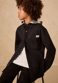 Camicia nera con bottoni e taschino sul petto, indossata sopra una camicia bianca, con cappuccio a scacchi grigi e neri. Tessuto dalla texture liscia.