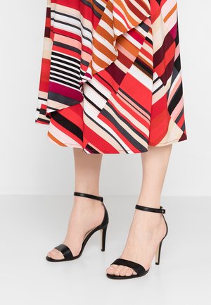 Jambes portant des sandales à talons hauts noires avec des brides à la cheville, et une jupe rayée colorée avec des motifs diagonaux rouges, orange, blancs, roses et noirs.