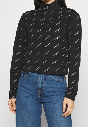 Femme portant un sweat-shirt zippé noir Calvin Klein avec logo imprimé et un jean bleu, debout devant un fond uni.