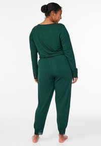 Zeleni dolg rokav kombinezon z čipkasto obrobo na manšetah in gležnjih. Mehki material, sproščen kroj in dizajn z zavijalnim hrbtnim delom. Minimalističen slog.