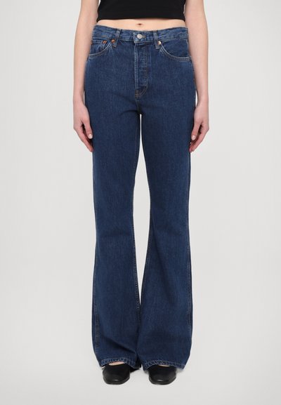 Mørkeblå denim flared jeans med høj talje, bæltestropper, fem lommer og orange syninger. Glat tekstur med en let glans.