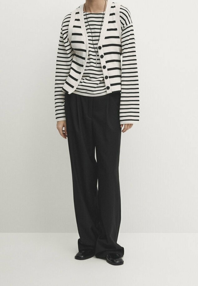 Personne portant un pantalon noir à jambes larges, des chaussures noires, une chemise à manches longues rayée horizontale noir et blanc et un gilet blanc boutonné avec des rayures.