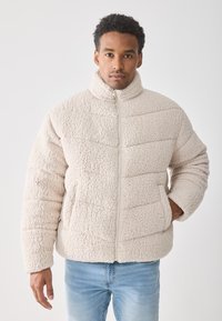 Muškarac u svijetlosmeđem teksturiranom zip-up jakni i plavim trapericama, stoji uz jednostavnu bijelu pozadinu, s jednom rukom u džepu.