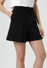 Shorts noirs sur mesure avec une large ceinture, des plis et des revers roulés. Confectionnés dans un tissu lisse, avec des poches latérales.