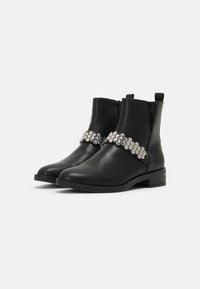Botines de cuero negro con tacones bajos, cremallera lateral y una correa decorada con hebillas hexagonales plateadas en la parte frontal.