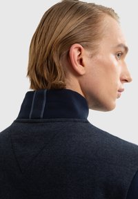 Jauns vīrietis ar blondiem matiem, tērpies tumši zila augsta apkaklīša džemperī, skatās pa labi pret gaismas pelēku fonu.