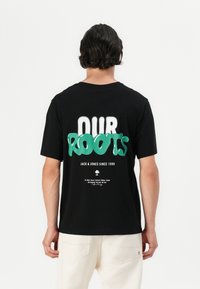Camiseta de algodón negra con grandes letras verdes que dicen "NUESTROS RAÍCES". Texto pequeño en la parte inferior que incluye "JACK & JONES DESDE 1990."