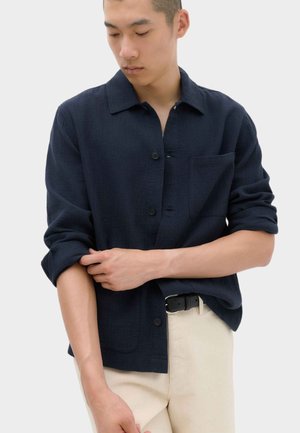 Mand justerer den opkrøllede ærme på en mørk navy knap-up skjorte, iført lyse beige bukser og et sort bælte, mod en ensfarvet baggrund.