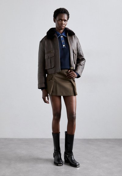 Giacca cropped in pelle marrone con colletto in pelliccia nera, camicia blu sotto, mini gonna oliva e stivali neri alti fino al ginocchio con borchie.