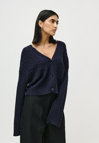 Kvinne med rett, mørkt hår iført marineblå, kabelstrikket knappet cardigan og svarte vide bukser, som står mot en enkel lyse bakgrunn.