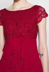 Czerwona sukienka z koronkowym gorsetem ozdobionym cekinami, ze falbaniastymi rękawami i zwiewną spódnicą z warstwowymi falbanami.