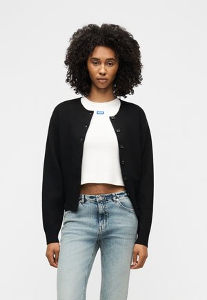 Žena s kučeravými vlasmi oblečená v čiernom zapínacom kardigane cez bielu skrátenú košeľu a svetlomodré džínsy, stojaca pred jednofarebným pozadím.