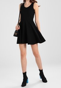 Frau in einem ärmellosen schwarzen Kleid mit weitem Schnitt, schwarzen Stiefeletten mit blauen Absätzen und einer kleinen schwarzen Handtasche mit Leopardenmuster-Details.