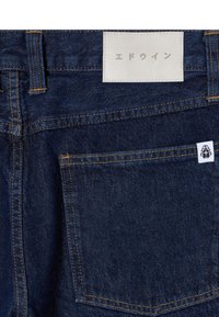 Jeans in denim blu scuro con taglio dritto, dotati di cuciture arancioni, un'etichetta in pelle con scritta in giapponese e una piccola etichetta con il marchio sulla tasca.