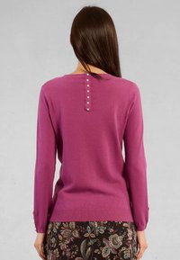 Donna con lunghi capelli castani che indossa un maglione viola a maniche lunghe con dettagli di bottoni perlacei sul retro e una gonna a fantasia floreale.