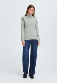 Hellgrüner Rollkragenpullover aus weichem Strickmaterial, kombiniert mit dunkelblauen, hoch geschnittenen, weiten Jeans und schwarzen Schuhen.
