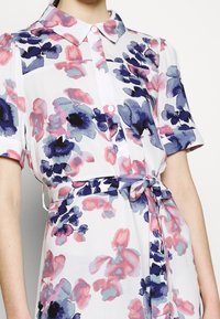 Abito floreale in tessuto leggero con motivi di fiori rosa, viola e blu. Include un collo a camicia e una cintura in vita legata.